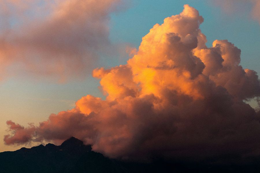 Een grote, dramatische wolkenformatie boven een silhouet van een bergkam bij zonsondergang, met warme tinten van oranje en roze in de lucht.