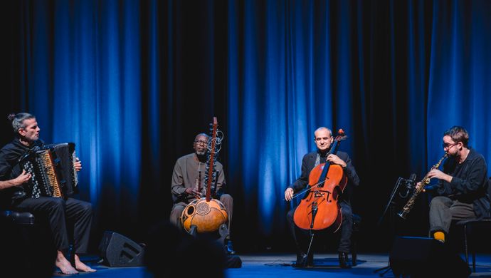 Vier muzikanten spelen in een intieme, blauw verlichte setting op accordeon, kora, cello en een klein snaarinstrument tijdens een sfeervol podiumoptreden.