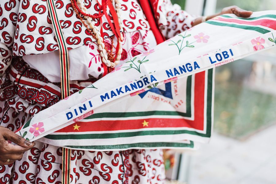 Persoon in traditionele kleding houdt een kleurrijke driehoekige sjaal vast waarop de tekst staat: "Ratu Makandre Tangi Libi".