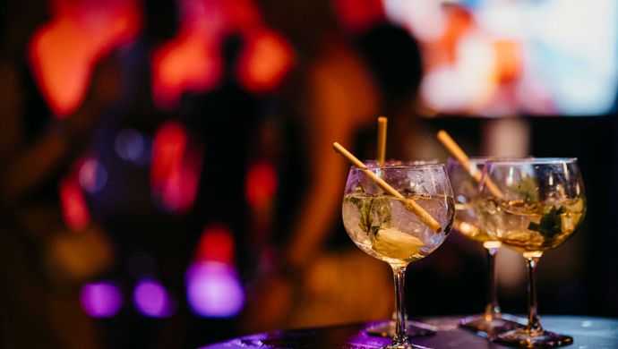 Two cocktail glasses with straws sit on a table during Disco Royale at de Doelen. In the background, partygoers and dancing silhouettes are visible in a colorfully lit space, creating a vibrant and festive atmosphere.