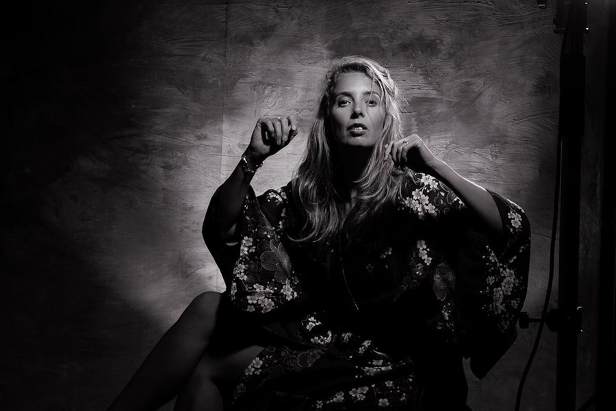 Zwart-wit portret van een vrouw met lang haar in een expressieve pose, gehuld in een volumineus kledingstuk met bloemenpatroon. Ze wordt van achteren belicht, waardoor een aura van licht haar omringt.