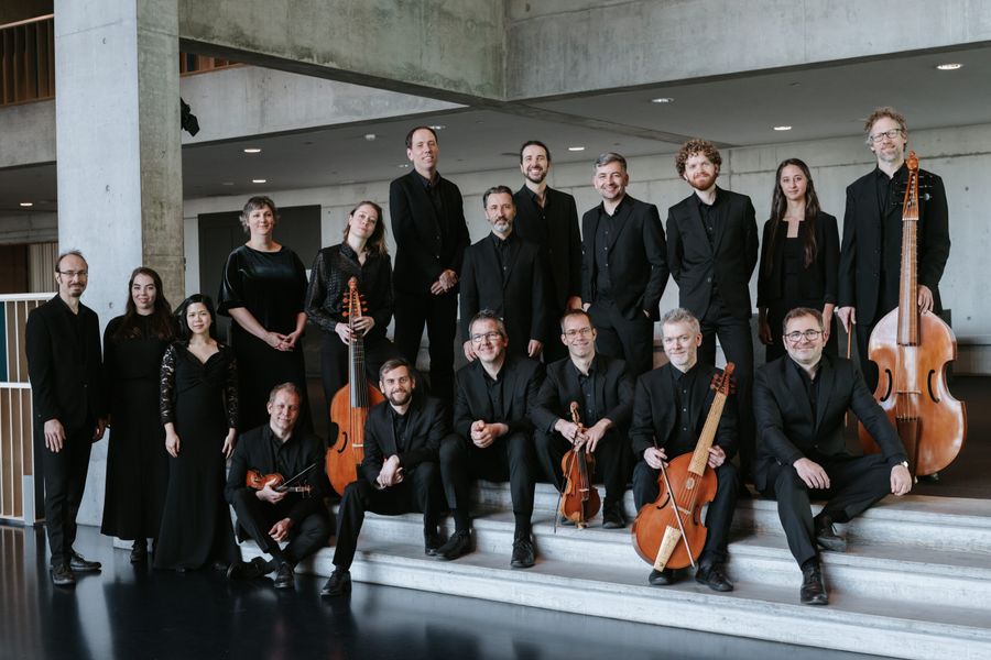 Large symphony orchestra dressed in black performs on a lit stage in a concert hall, featuring strings, winds, and percussion. A grand pipe organ is visible in the background, and the audience watches attentively.