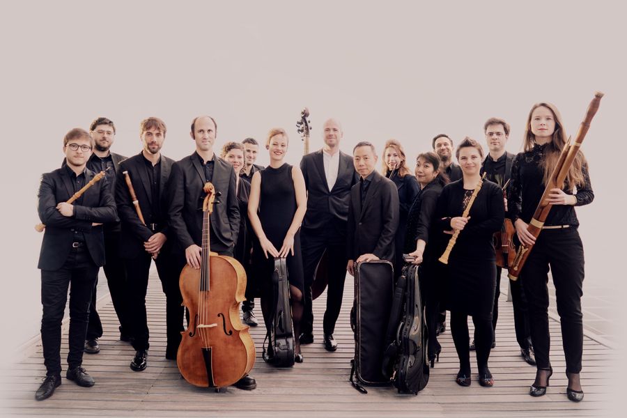 Een groep van veertien musici poseert in een lichte studio-omgeving, gekleed in zwarte kleding. Ze houden verschillende instrumenten vast, waaronder een cello, dwarsfluit, contrabas, trompet en hob