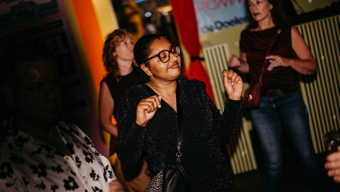 A woman dances with a smile on her face during Disco Royale at de Doelen. She wears a sparkly black outfit and stands in a colorfully lit space, with other partygoers in the background.