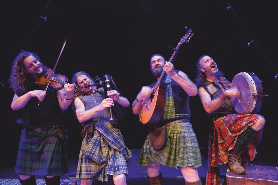 Vier muzikanten in Schotse kilts spelen energiek viool, gitaar en percussie op een podium, met expressieve houdingen en uitbundige gezichtsuitdrukkingen.
