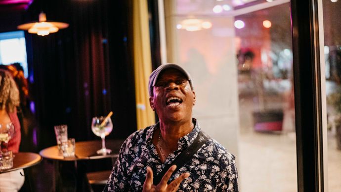 A man wearing a cap and a patterned shirt looks up laughing during Disco Royale at de Doelen. He stands near a window by the bar, with a cocktail glass and warm lighting adding to the festive setting.