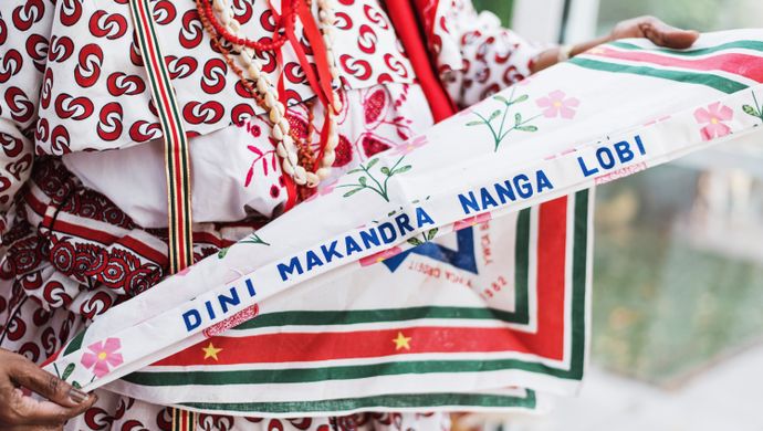 Persoon in traditionele kleding houdt een kleurrijke driehoekige sjaal vast waarop de tekst staat: "Ratu Makandre Tangi Libi".