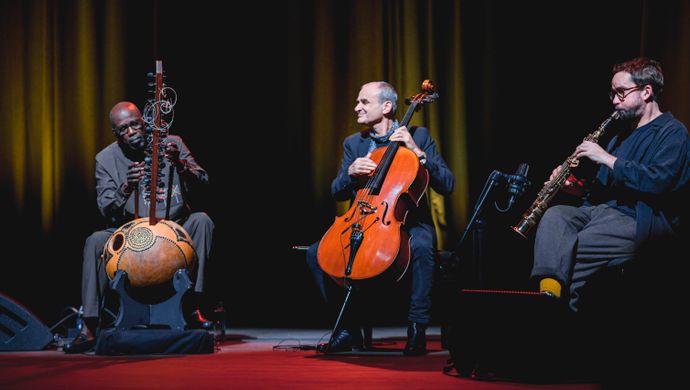 Vier muzikanten spelen in een intieme, geel verlichte setting op accordeon, kora, cello en een klein snaarinstrument tijdens een sfeervol podiumoptreden.