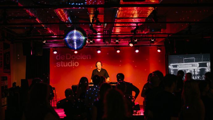 The audience dances in front of the stage during Disco Royale at de Doelen Studio. The space is lit in red and purple, with a disco ball overhead and a DJ performing beneath the de Doelen Studio logo. On the right, a screen displays moving visuals.