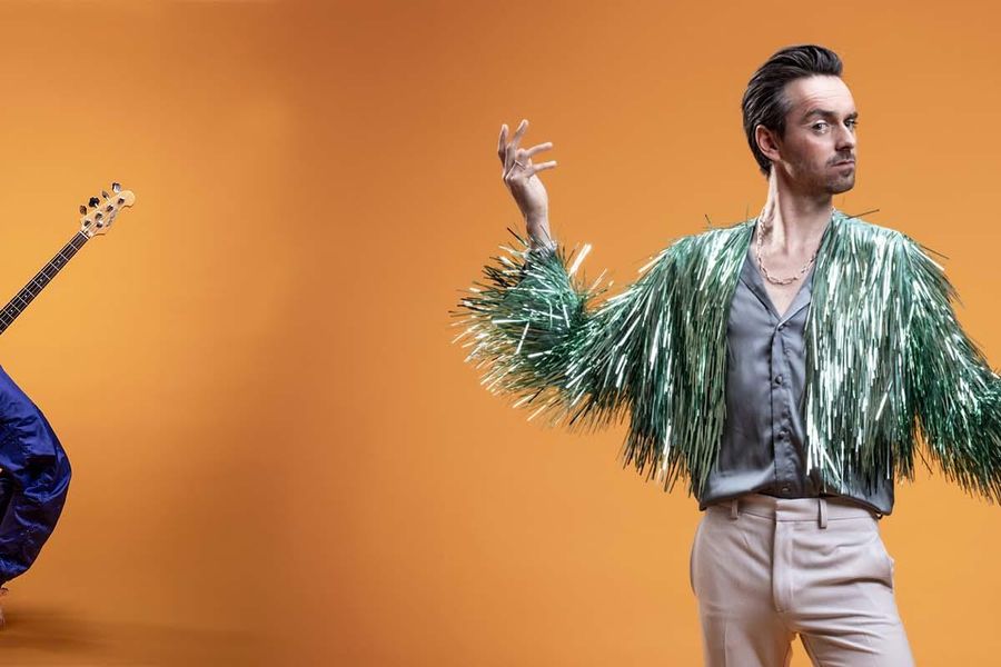 Twee muzikanten op een warme oranjebruine achtergrond. Links zit een vrouw met een elektrische gitaar, gekleed in een kleurrijk topje en blauwe broek. Rechts staat een man met een viool in de hand en een expressieve houding, gekleed in een opvallend groen met wit gewaad.