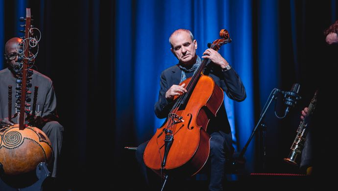 Three musicians play the kora, cello, and a small string instrument in an intimate, blue-lit setting during an atmospheric stage performance.