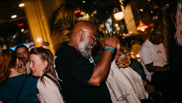 Partygoers dance close together during Disco Royale at de Doelen. In the foreground, an energetic man with a grey beard is dancing, surrounded by laughing people in a busy, vibrant, and festive atmosphere.