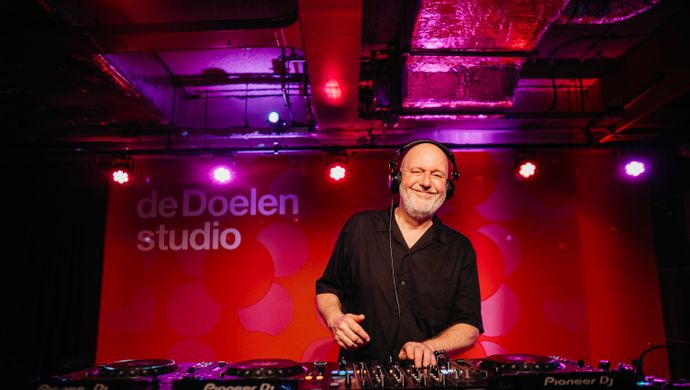 The DJ smiles while performing during Disco Royale at de Doelen Studio. He stands behind a DJ booth in a room lit with purple and pink lights. On the wall behind him, the logo of de Doelen Studio is visible.