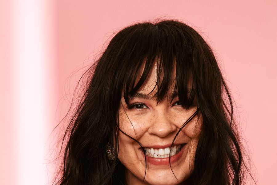 Vrouw met lang donker haar en een brede lach, gekleed in een felroze outfit, poseert voor een roze achtergrond met wapperend haar.