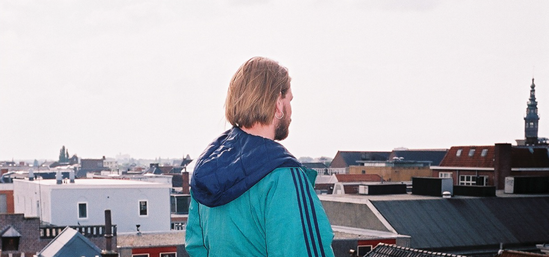 Foto van een man in een blauwe jas, die buiten op een dak wegkijkt van de camera. 