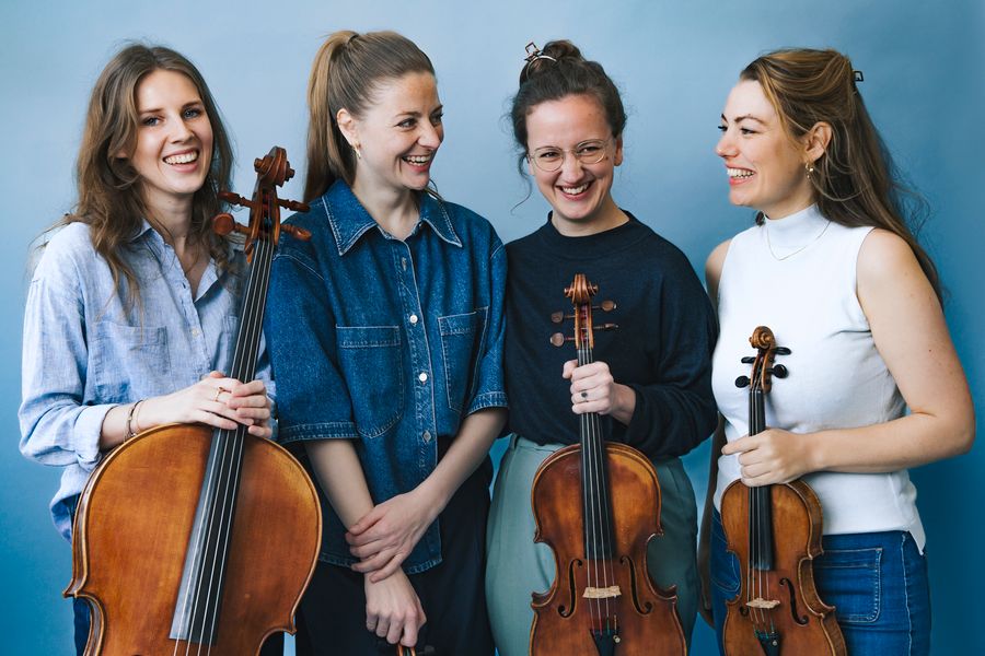 Vier vrouwelijke muzikanten lachen samen voor een blauwe achtergrond. Ze houden strijkinstrumenten vast: twee violen, een altviool en een cello. Hun houding is ontspannen en vriendschappelijk, wat een gevoel van verbondenheid en plezier uitstraalt.