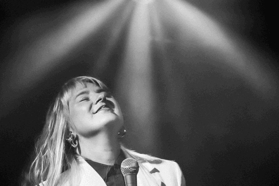 Zangeres in een wit jasje staat onder een spotlight, glimlachend met gesloten ogen en een microfoon in de hand, terwijl het licht haar van boven verlicht.