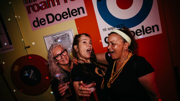 Three women joyfully pose together  for the camera during Disco Royale at de Doelen. They are holding drinks and standing close in front of a colorful background featuring retro-style posters and red disco lighting. A playful, energetic vibe captures the festive spirit of the night.