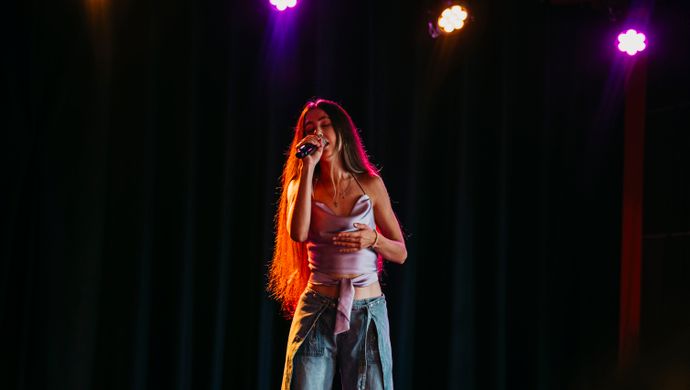 Een jonge zangeres staat solo op het podium tijdens de Vocal Night van de Jordan Roy Vocal Academy in Studio 1 van de Doelen. Ze zingt vol overgave onder warme theaterlichten, tegen een donkere achtergrond.