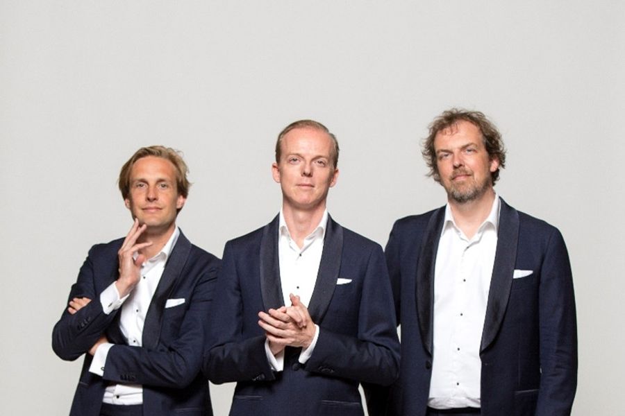 Drie mannen in nette pakken poseren voor een lichtgrijze achtergrond. Ze staan naast elkaar, lachen vriendelijk en kijken in de camera.