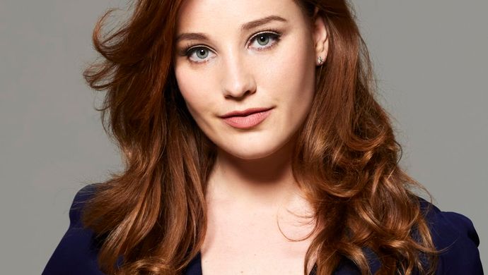 Woman with long, wavy brown hair wearing a dark blue V-neck outfit, looking confidently at the camera against a grey background.