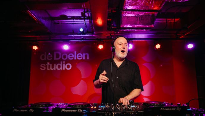 The DJ stands behind his turntables during Disco Royale at de Doelen Studio. He is dressed in black and lit by purple and pink spotlights, with a red wall in the background displaying the words 'de Doelen studio'.