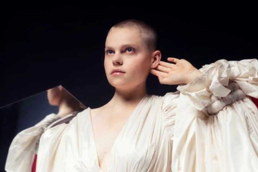 Een persoon met een kaalgeschoren hoofd draagt een dramatische, wijdvallende witte outfit met rode accenten. Ze kijken zelfverzekerd voor zich uit en houden hun handen bij hun oren. De achtergrond is donker, wat de theatrale belichting versterkt.