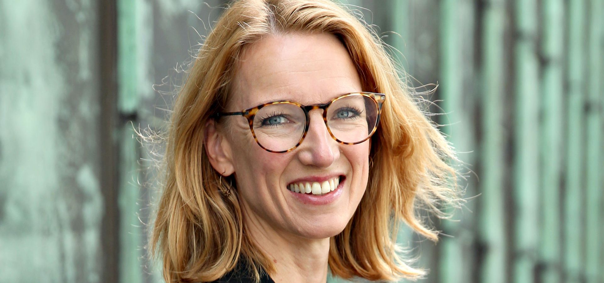 Woman with blonde hair and glasses smiling at the camera, wearing a black blazer, posing outdoors in front of a weathered metal wall.