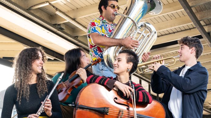 Vijf jonge muzikanten spelen klassieke instrumenten op een stedelijke buitenlocatie onder een viaduct. Ze bespelen onder andere viool, cello, klarinet en tuba, en hebben een energieke en vrolijke uitstraling.