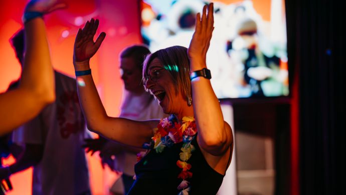 A joyful woman dances with her arms raised during Disco Royale at de Doelen. She wears a colorful Hawaiian lei and a black outfit, laughing exuberantly as disco visuals play on a screen behind her. Red and purple lighting enhance the festive atmosphere.