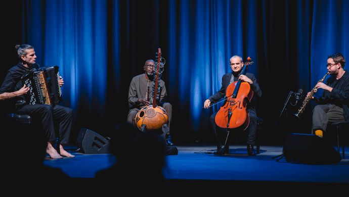 Vier muzikanten spelen in een intieme, blauw verlichte setting op accordeon, kora, cello en een klein snaarinstrument tijdens een sfeervol podiumoptreden.