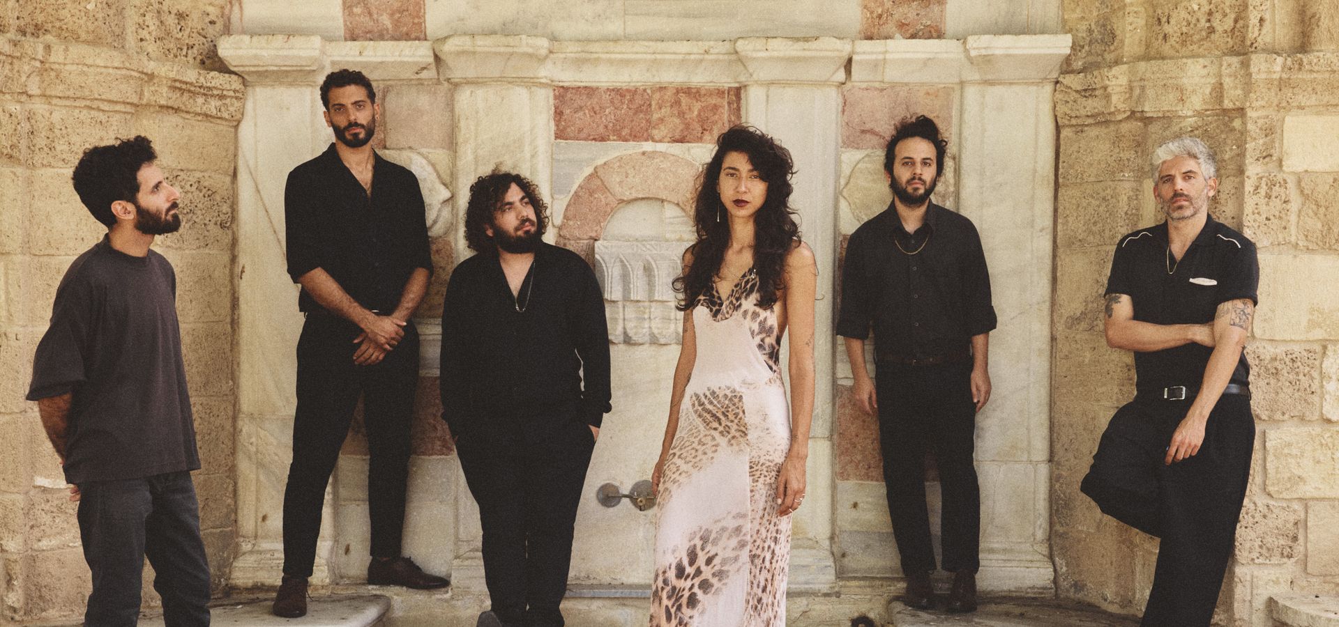 Zes bandleden staan bij een stenen muur. Eén persoon draagt een lichte jurk met patroon, de anderen zijn in donkere kleding gekleed.