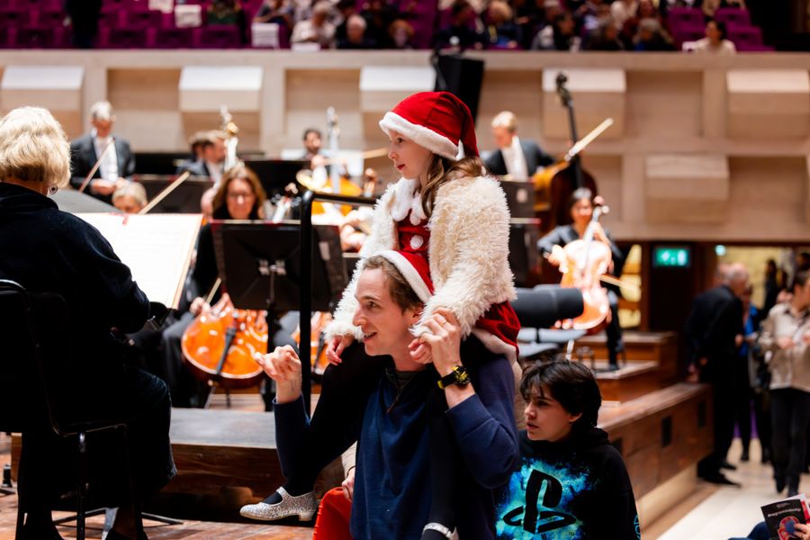 Een vader met twee kinderen in een concertzaal, waarvan één op zijn schouders zit met een kerstmuts op. Op de achtergrond repeteert een orkest, en de sfeer is feestelijk en warm.