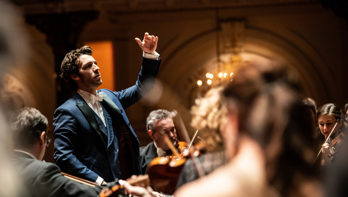 Conductor in a blue suit leads an orchestra during a performance in a classical concert hall with warm lighting and elegant architecture.