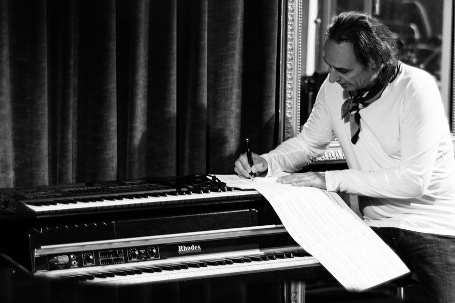 Een man in een witte blouse schrijft muzieknoten op een groot vel papier dat over een elektrische piano is uitgespreid, in een intieme, studiovormige omgeving.