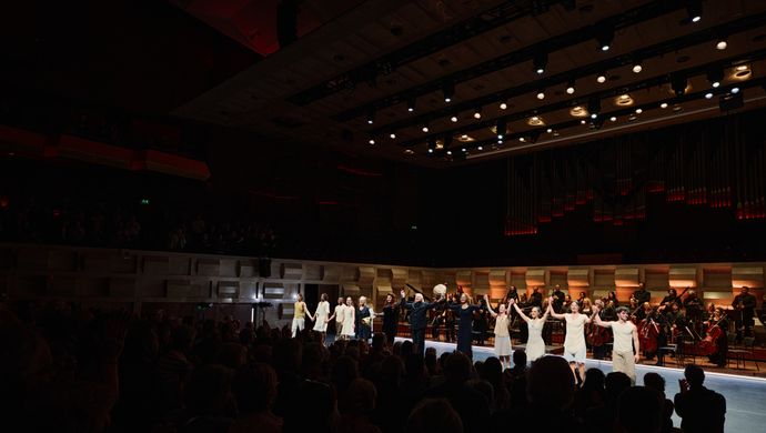 Het volledige gezelschap van Conny Janssen Danst, samen met dirigent Conrad van Alphen en sopraan Ewa Tracz, buigen hand in hand op het podium van de Grote Zaal in de Doelen tijdens het slotapplaus van Górecki’s Symphony of Sorrowful Songs, uitgevoerd door Sinfonia Rotterdam. Op de achtergrond applaudisseren de musici van het orkest en het publiek in een uitverkochte zaal.