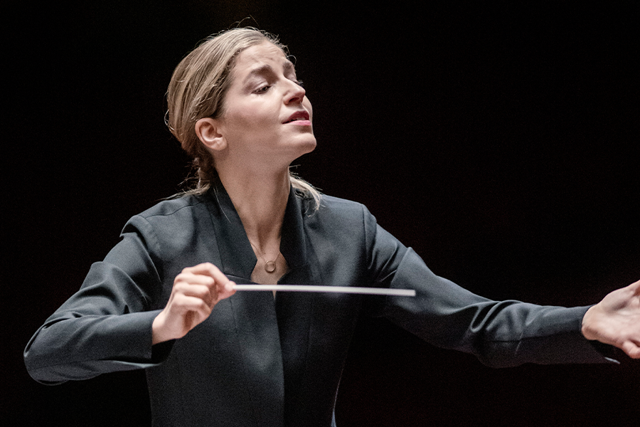 Dirigente in een zwarte outfit leidt een orkest met expressieve handgebaren, geconcentreerd kijkend tegen een donkere achtergrond.