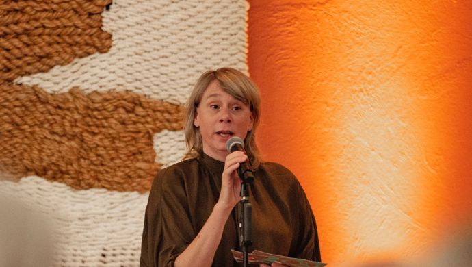 Vrouw in bruin shirt praat tegen het publiek in een microfoon