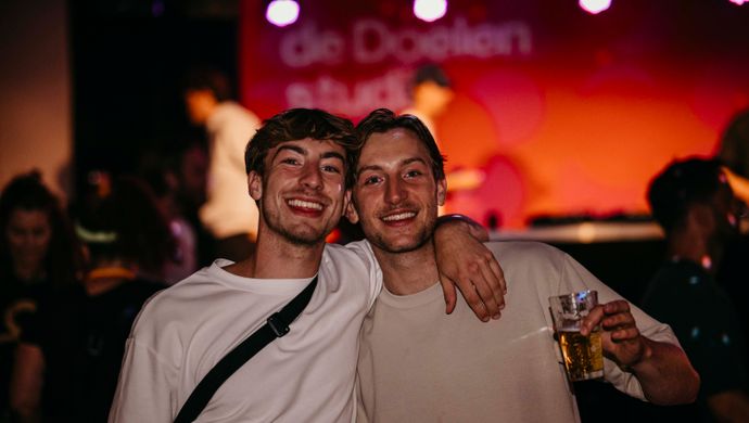 Two young men pose smiling with their arms around each other during Disco Royale at de Doelen Studio. They are holding drinks, with a red-lit stage in the background and a festive atmosphere all around.
