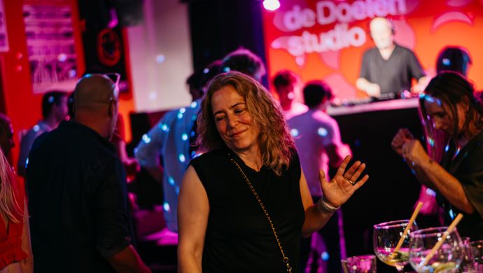 A woman dances casually on the dance floor during Disco Royale at de Doelen Studio. In the background, a DJ is playing music and other partygoers are visible, surrounded by colorful lights and an energetic atmosphere.