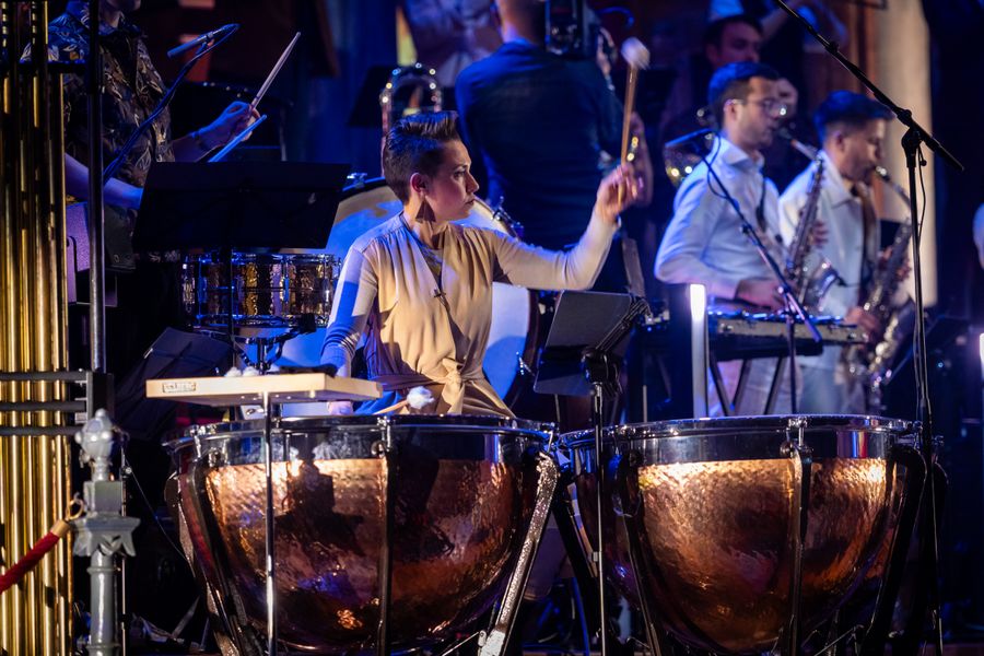 Een slagwerker speelt op pauken tijdens een concert, gefocust en energiek. Andere musici en instrumenten zijn zichtbaar op de achtergrond in een sfeervol verlichte concertzaal.