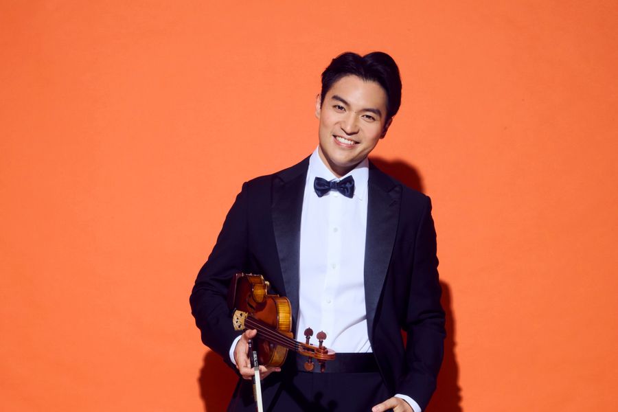 Een man in smoking met een vlinderdas poseert met een viool in zijn hand tegen een feloranje achtergrond.