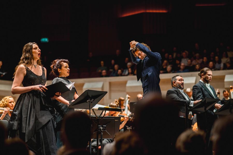 Dirigent leidt een orkest met twee klassieke zangers op de voorgrond tijdens een concert; de zangers houden partituren vast en zingen voor een publiek in een concertzaal.