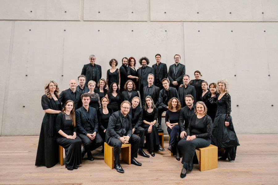 Groepsfoto van ongeveer 25 mensen, gekleed in zwart, poserend in twee rijen voor een lichte betonnen muur; sommigen zitten op houten blokken, anderen staan achter hen.