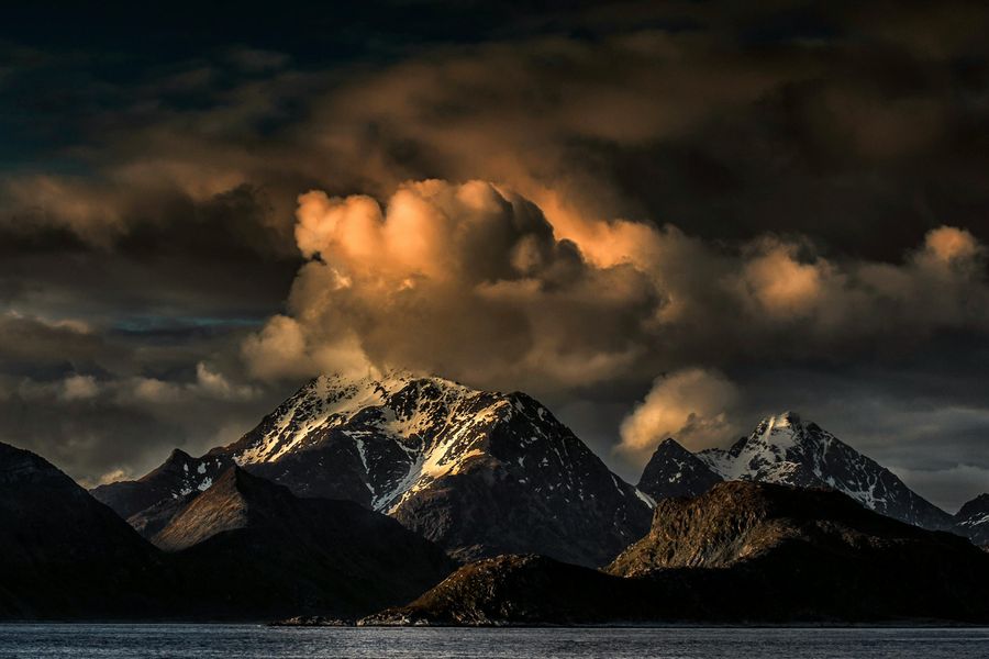 Een indrukwekkend berglandschap bij zonsondergang, met besneeuwde toppen die oplichten in het warme, oranje licht van dramatische wolken. De voorgrond toont een rustig meer dat het schouwspel weerspiegelt.