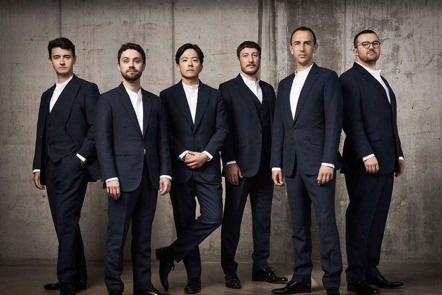 Groepsfoto van zes mannen in donkere kostuums met witte overhemden, die in een nette rij poseren tegen een betonnen muur. Ze kijken serieus en professioneel naar de camera.