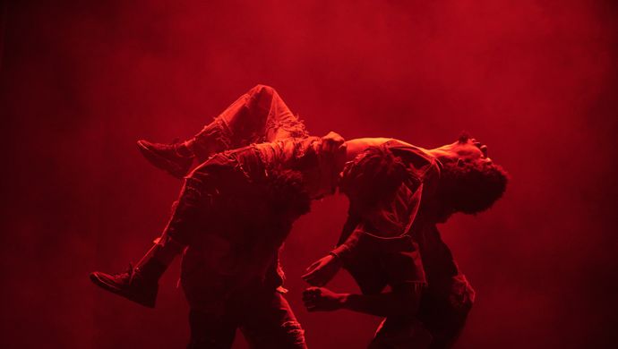 Twee dansers voeren een expressieve choreografie uit in rood toneellicht. Eén danser houdt de ander dramatisch gebogen over de schouders, terwijl rook of mist de achtergrond vult.