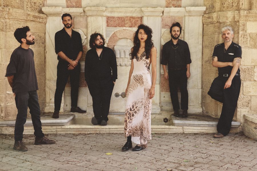 Zes bandleden staan bij een stenen muur. Eén persoon draagt een lichte jurk met patroon, de anderen zijn in donkere kleding gekleed.