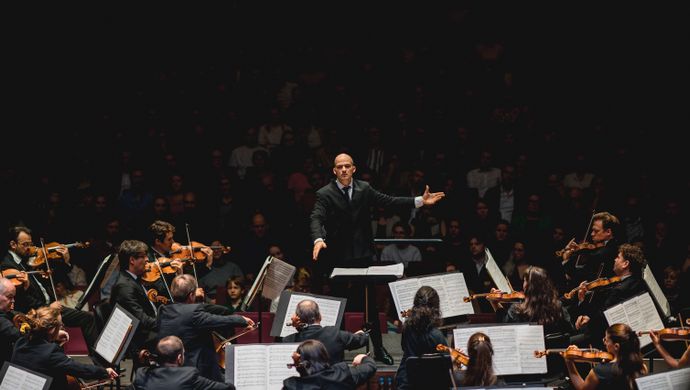 Orkestleden spelen en kijken naar de dirigent terwijl het publiek toekijkt