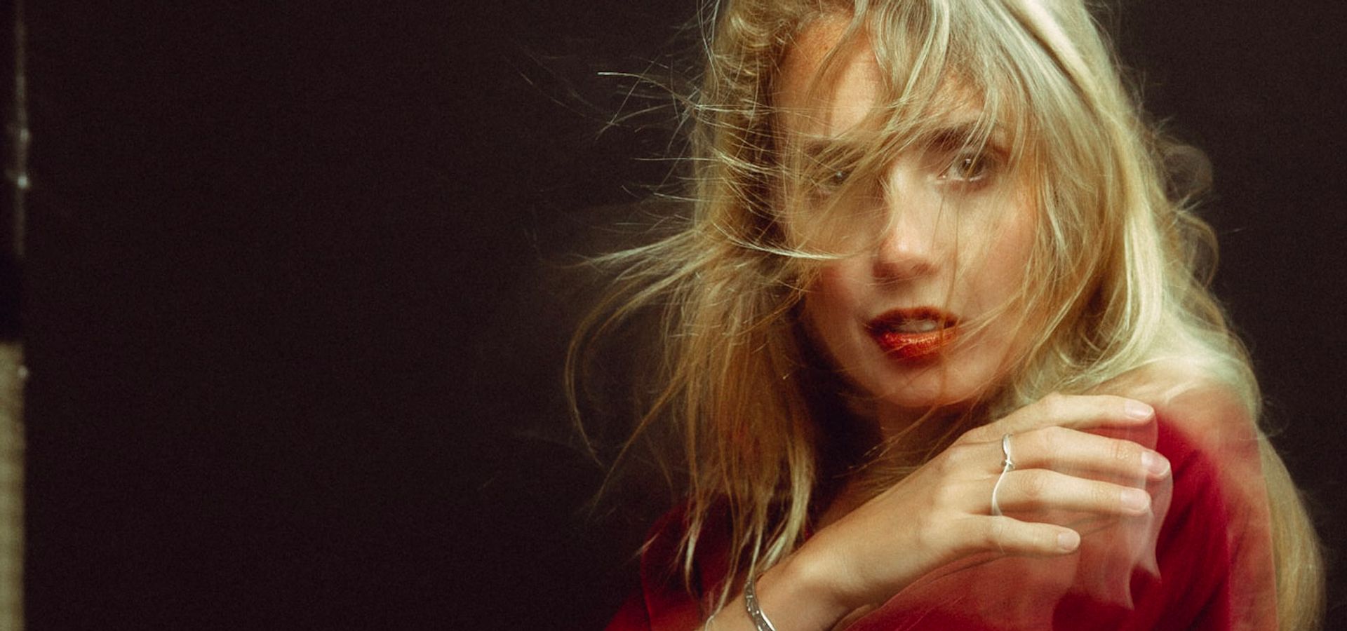 Woman with long blonde hair wearing a red velvet dress sits on a dark leather couch, gazing at the camera with a confident, slightly mysterious expression.